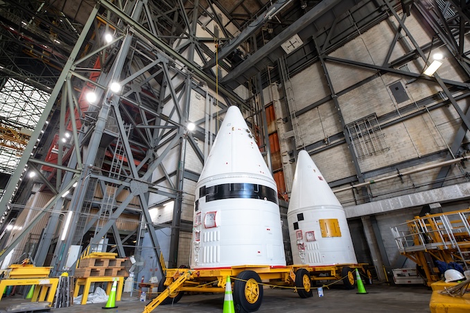 Two large white rocket-shaped payload fairings sit on yellow transport carts inside a tall industrial facility. The building’s interior is filled with steel beams, platforms, and bright overhead lights.